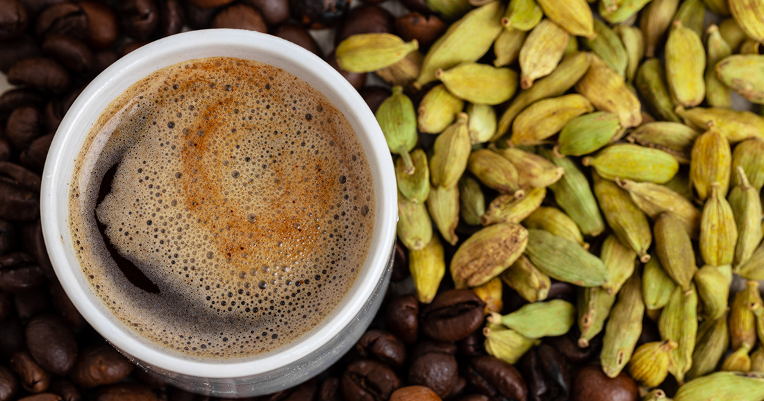 Freshly brewed cup of coffee surrounded by cardamom pods and roasted coffee beans.