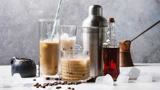 Iced coffee cocktail setup – Assorted iced coffee cocktails in glasses of various sizes, with ice cubes, coffee beans, and a cocktail shaker arranged on a white surface.