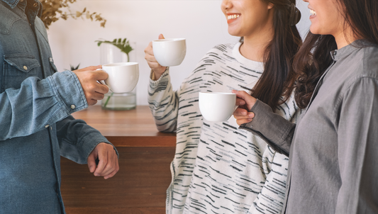 Group of friends smiling and holding white coffee mugs, enjoying a warm coffee break together indoors.