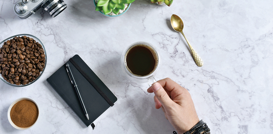 Top view of a fresh black coffee held by hand on a marble desk, surrounded by coffee beans, a notebook, and workspace essentials for a focused morning routine.