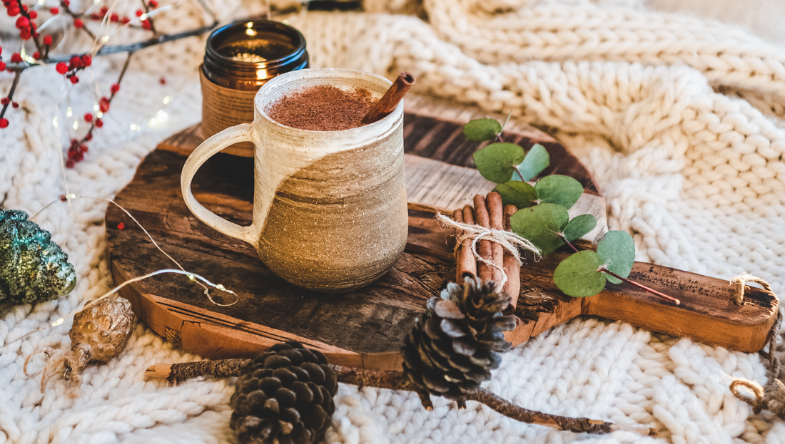 A cozy winter scene featuring a rustic ceramic mug filled with spiced coffee or hot chocolate, topped with cinnamon and placed on a wooden board surrounded by pinecones, eucalyptus leaves, cinnamon sticks, and warm knitted fabric.