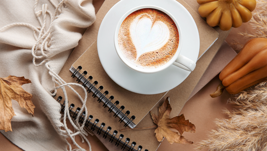 A warm cup of coffee with heart-shaped foam surrounded by notebooks, autumn leaves, and soft beige textures — the perfect cozy fall morning.