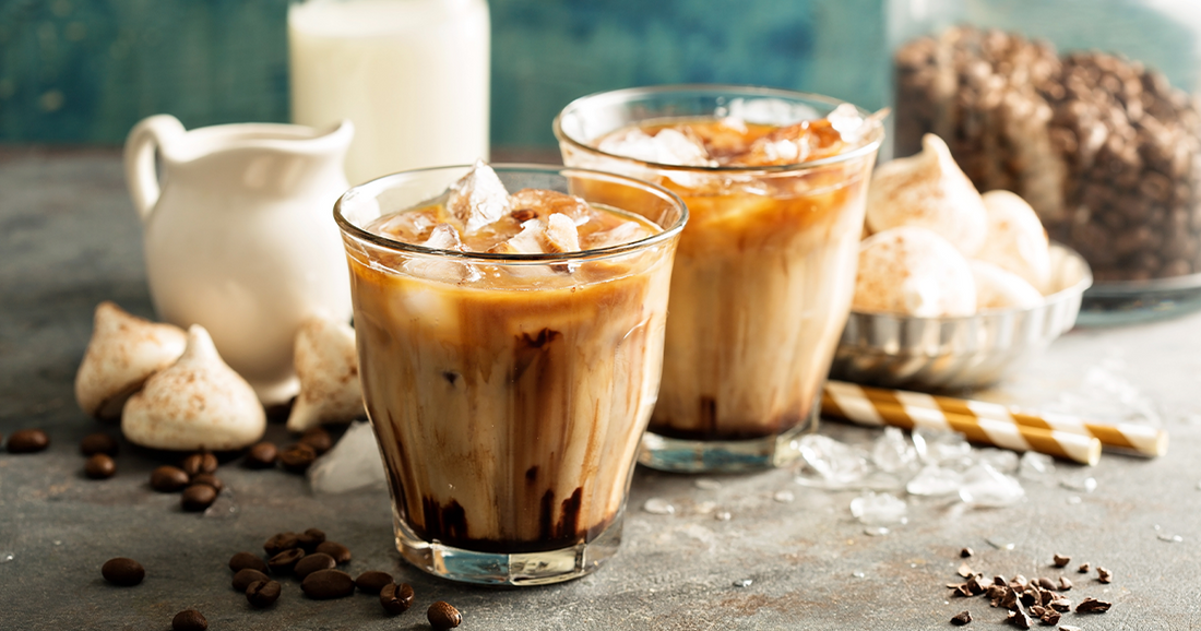 Iced coffee with milk served in clear glasses, topped with ice, surrounded by coffee beans, meringue cookies, and milk jug.