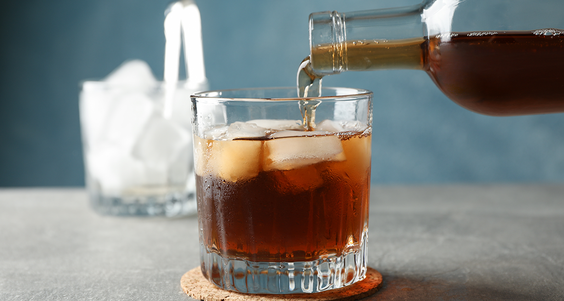 Cold brew coffee being poured over ice in a clear glass on a gray background.