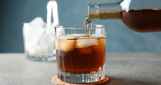 Cold brew coffee being poured over ice in a clear glass on a gray background.