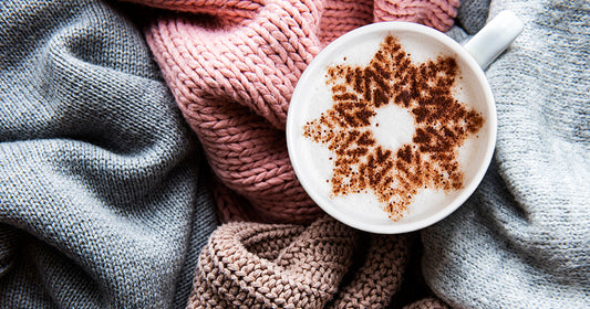Cup of cappuccino with snowflake pattern on frothy milk resting on cozy knitted blankets in soft winter tones.